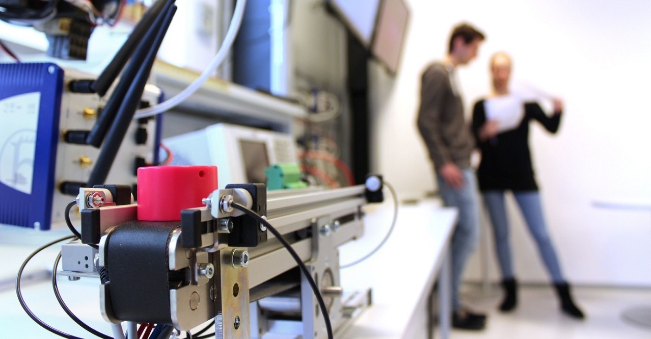 Industrielle Automatisierungstechnik mit vernetzten Steuerungssystemen im Fokus. Zwei Ingenieure diskutieren im Hintergrund über IT-Sicherheitsaspekte in der smarten Fertigung.