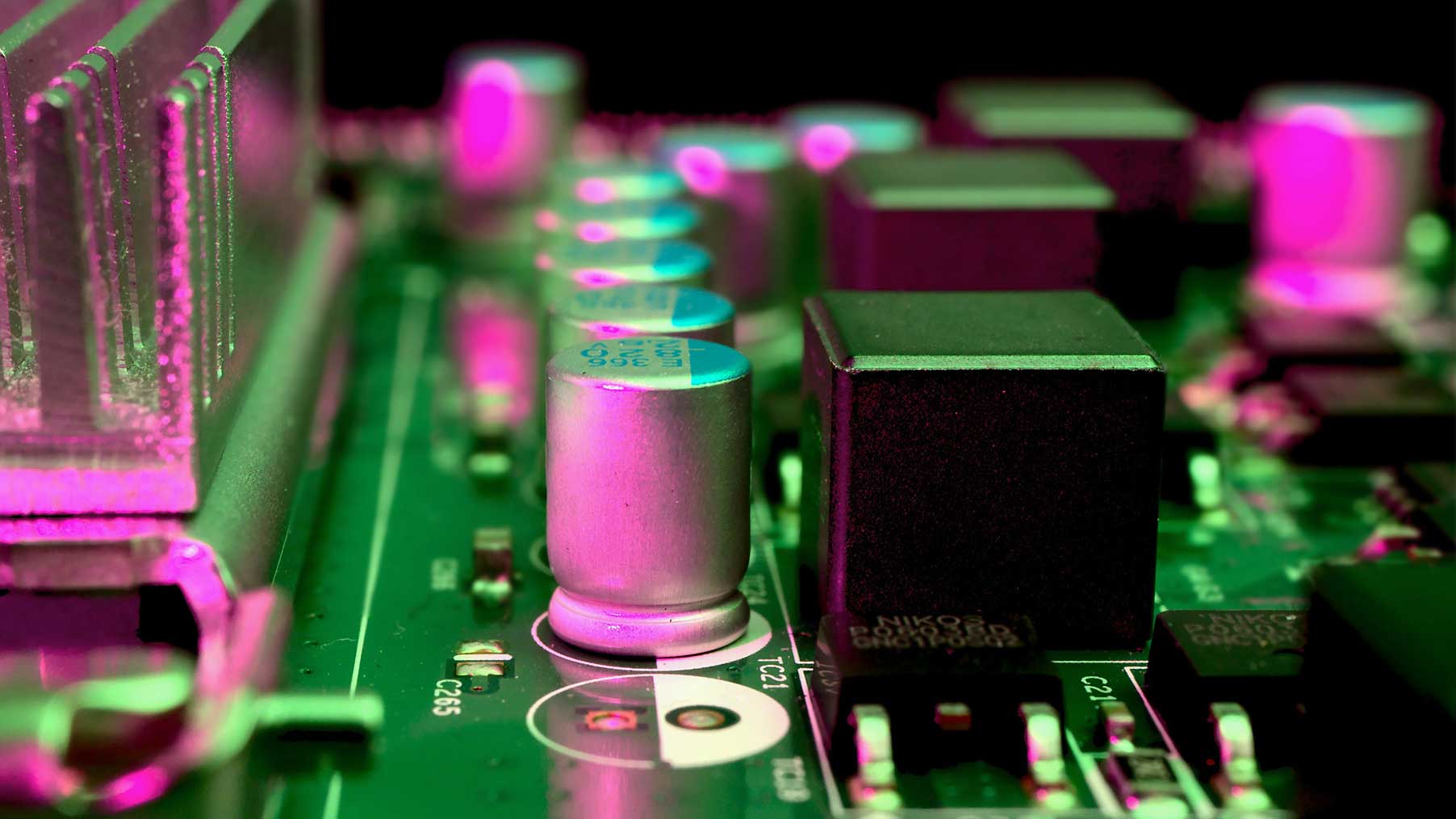 Nahaufnahme einer Leiterplatte mit elektronischen Komponenten wie Kondensatoren und Kühlkörpern, beleuchtet in Neonfarben.