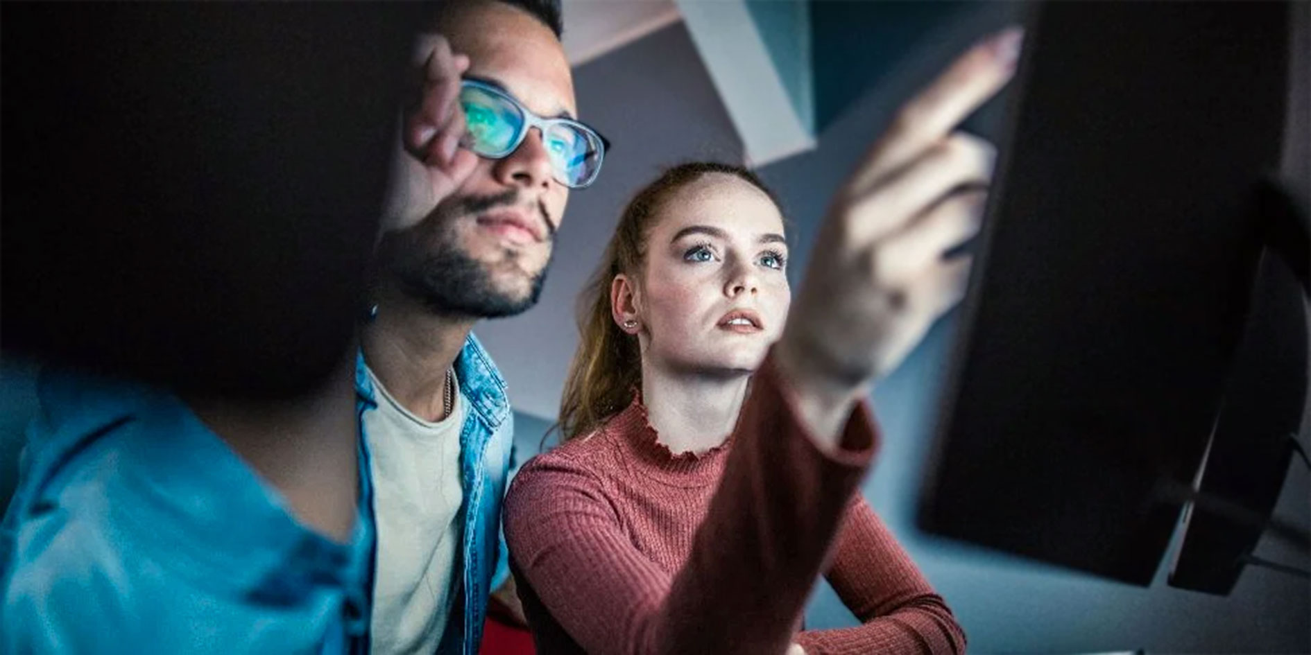Zwei IT-Spezialisten analysieren Daten auf einem Computerbildschirm. Eine Frau in einem roten Pullover zeigt konzentriert auf den Monitor, während ein Mann mit Brille aufmerksam zusieht.